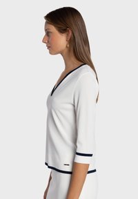 Haut blanc à col en V avec manches trois-quarts ornées de rayures bleu marine aux poignets et à l'ourlet, confectionné dans un tissu lisse, présentant une coupe décontractée.