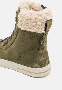 Botín de ante verde oliva de caña alta con un suave puño de piel de color beige, con una etiqueta de logo negra y una delgada suela de goma blanca con franjas beige.