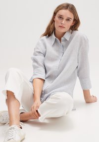 Light blue striped button-up shirt with long sleeves, paired with white cropped pants, and light sneakers on a neutral background.