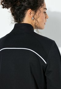 Schwarzer Pullover mit hohem Kragen, der einen weißen Akzentstreifen auf der Rückseite zeigt. Der Stoff wirkt weich und strukturiert, was Tiefe verleiht.