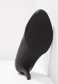 Chaussure à talon haut en cuir noir avec un bout pointu et un talon aiguille fin, présentant une texture lisse et un motif floral subtil sur la semelle.