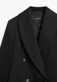 Černý dvouřadý blazer se šálovým límcem, vyrobený z texturované látky, s dvěma černými knoflíky a vnitřní štítkem.