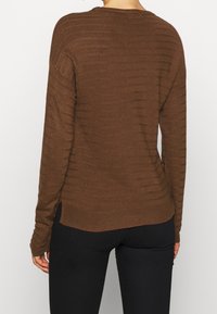 Persona che indossa un maglione lungo marrone a maniche lunghe con texture e pantaloni neri, vista da dietro su uno sfondo bianco semplice.