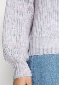 Gros plan d'une personne portant un pull tricoté violet clair avec des poignets et un ourlet côtelés, associé à un jean en denim bleu.