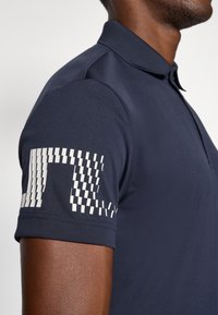 Marineblå polo-shirt, kortermet, med et hvitt geometrisk mønster på venstre arm. Glatt stoff med standard krage og tettsittende design.