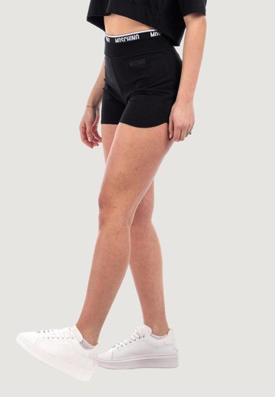 Pantaloni corti sportivi neri con una vita alta che presenta il marchio Moschino, abbinati a sneakers bianche. Il tessuto appare liscio e aderente.