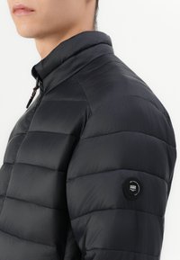 Black puffer jacket with a high collar, featuring horizontal stitching, a round logo patch on the sleeve, and a smooth, shiny texture.