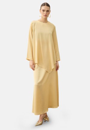 Donna che indossa un lungo vestito giallo chiaro con orlo asimmetrico e maniche lunghe, abbinato a décolleté dorate con punta e orecchini a cerchio.