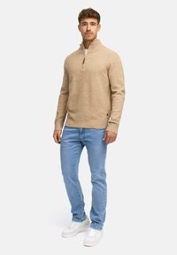 Hellbrauner Zip-Pullover aus weichem Material, kombiniert mit hellblauen Jeans und weißen Sneakern. Der Pullover hat gerippte Bündchen und einen Saum.