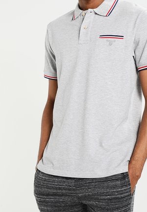 Man wearing light gray polo shirt with red, white, and blue striped collar and sleeve edges, paired with dark gray textured pants.