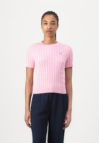 Pull en tricot rose à manches courtes avec un motif en relief de type cable et un ourlet côtelé, associé à un pantalon bleu marine. Présente un petit logo sur la poitrine.
