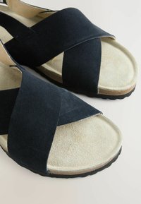 Sandales en suède noir avec de larges sangles croisées, une semelle intérieure beige douce et une semelle en caoutchouc texturé avec un léger talon.