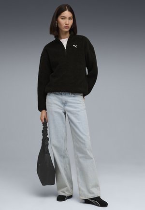 Sort fleece-hoodie med halv lynlås og hvidt logo, parret med lyseblå brede bukser og sorte slip-on sko, mens der holdes en sort taske.