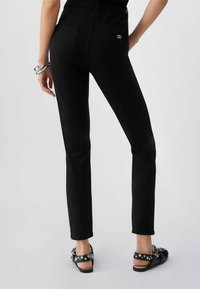 Persona que lleva pantalones ajustados negros con bolsillos traseros, un pequeño detalle de logotipo dorado, pulseras plateadas en una muñeca y sandalias negras con tachuelas.