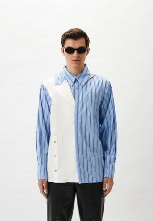 Gestreiftes Hemd in Blau und Weiß mit asymmetrischem Design, das ein solides weißes Panel, Knopfdetails und lange Ärmel aufweist. Glatte Stofftextur.