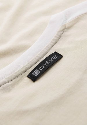 Vaalean beige puuvillapaita, jossa on valkoinen pyöreä kaula-aukko ja musta kangastunniste, jossa on "ombre"-logo tikattuna sivussa.