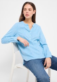 Hellblause Hemdbluse mit kleinen weißen Mustern, Rundhalsausschnitt und langen Ärmeln, aus einem weichen, leichten Stoff gefertigt.