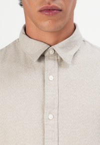 Homem com uma camisa de botão de cor bege clara com manchas, focando no colarinho e na parte superior do peito.