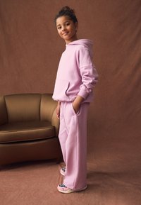 Sudadera con capucha de color rosa claro y pantalones deportivos a juego hechos de tela suave; presenta un bolsillo frontal y un ajuste relajado. Acompañado de pantuflas con un estampado divertido.