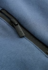 Tessuto blu con una cerniera dotata di un tirante nero. Le cuciture visibili e la sottile trama del tessuto evidenziano i dettagli del design.