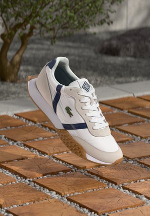 Witte sneaker met marineblauwe accenten en een getextureerde gumzool. Voorzien van een suede neuskap en geborduurd logo aan de zijkant. Vetersluitingen.