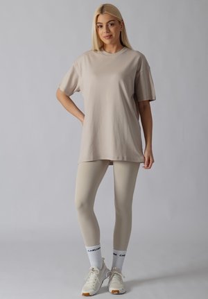 Femme aux longs cheveux blonds portant un t-shirt beige oversize, des leggings assortis, des chaussettes blanches de marque et des baskets blanches, debout sur fond gris.