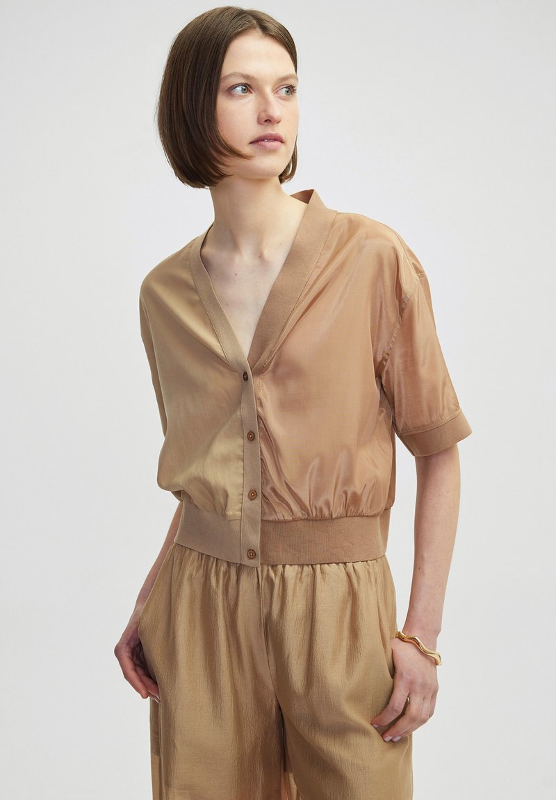 Kurzärmliges, v-ausschnittenes Shirt in zwei Beigetönen, mit Knopfverschluss und geripptem Saum. Glattes, glänzendes Material mit lockerer Passform.