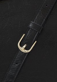 Gros plan sur un sac en cuir noir à surface texturée, avec une boucle en métal doré et une sangle, mettant en valeur le design épuré et la qualité du matériau.