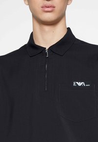 Černé polo triko s límcem na zip, kapsou na hrudi s bílým logem a hladkou texturou.