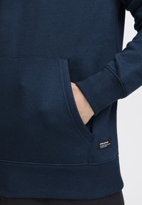 Felpa blu navy con una grande tasca frontale, polsini a costina e fondo a costina. Presenta un piccolo etichetta di marca nella parte inferiore sinistra. Tessuto morbido.