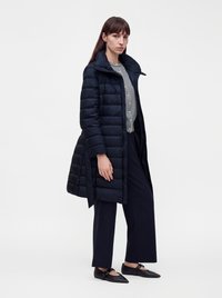 Femme portant un long manteau noir matelassé par-dessus une chemise rayée, un pantalon large noir et des ballerines Mary Jane noires, debout devant un fond blanc.