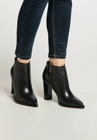 Sorte læder ankelstøvler med spids tå, høj blokhæl og lynlås i siden. Båret sammen med mørke jeans, der viser en glat og elegant tekstur.