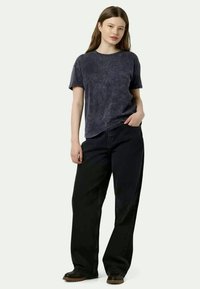 Νεαρή γυναίκα που στέκεται με το ένα χέρι στην τσέπη, φορώντας ένα σκούρο τζιν μπλουζάκι, χαλαρούς μαύρους παντελόνες και μαύρα παπούτσια σε απλό φόντο.