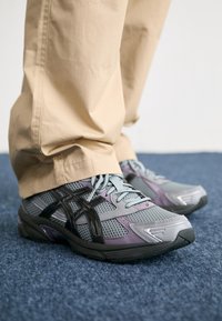 Persoon in beige broek en grijs-paarse sportchoenen met zwarte strepen, staande op een blauwe tapijt.