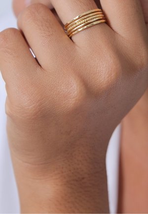 Gros plan sur une main à la carnation moyenne portant quatre fines bagues dorées texturées empilées sur le doigt du milieu.