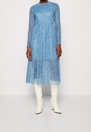 Blaues Midi-Kleid mit langen Ärmeln, floralem Muster und transparentem Überzug, kombiniert mit kniehohen weißen Stiefeln mit Absatz an einer stehenden Person.