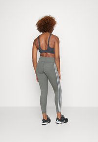 Soutien-gorge de sport gris avec bretelles réglables, associé à un legging vert à taille haute et texturé, présentant trois rayures blanches sur le côté et des chaussures de sport noires.