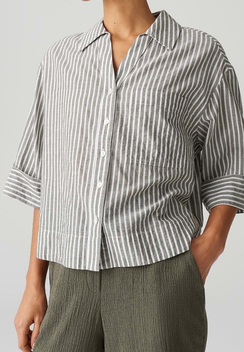 Femme portant une chemise ample à rayures verticales grises et blanches, boutonnée, à manches trois-quarts, et un pantalon texturé vert olive.