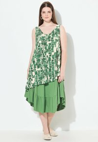 El vestido sin mangas presenta un patrón geométrico verde sobre un fondo marfil, con una falda sólida verde y fluida. Textura suave y caída.