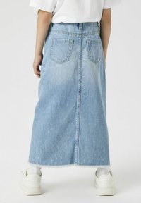 Helles blaue Denim-Maxi-Rock mit ausgefranstem Saum, zwei hinteren Taschen und geradem Schnitt. Getragen mit weißen Sneakers und sichtbaren Socken.