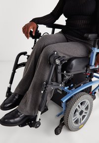 Calças cinza risca de giz cobrem as pernas de uma pessoa numa cadeira de rodas preta com detalhes azuis, apresentando apoios de braço pretos e uma roda prateada.