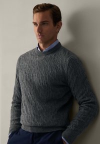 Grauer Strickpullover mit Kabelmuster, gerippten Bündchen und Saum. Getragen über einem hellblauen Hemd mit Kragen. Der Stoff wirkt weich.
