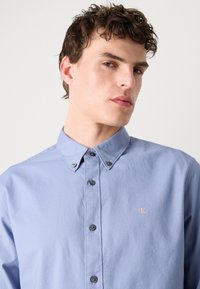 Camisa azul clara com botão e colarinho, apresentando um discreto logotipo CK em dourado e botões escuros na frente para fecho.