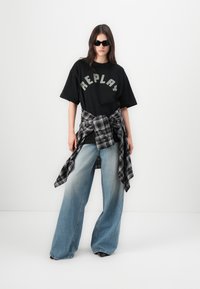 Svart T-skjorte med "REPLAY" logo, baggy blå jeans, og en svart og grå rutete skjorte knyttet rundt livet. Modell iført solbriller.