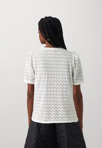 Hvit strikket topp med korte puff-ermer, med et teksturert mønster av skjellformer. Baksiden har et solid, sømløst design.