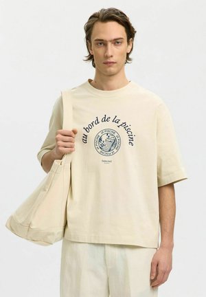 Fiatal férfi krémszínű pólóban, francia szöveg és grafika, a vállán tart egy hozzá illő krémszínű szövettáskát, egyszínű háttér előtt.