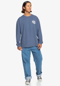 Niebieska bluzka z długim rękawem, okrągłym dekoltem i logo na lewej piersi, zestawiona z jasnoniebieskimi luźnymi dżinsami oraz czarno-białymi sneakersami.