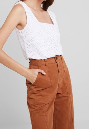 Kobieta ubrana w białą, bezrękawnikową bluzkę z ażurowym wzorem i wysokim stanem, rdzawobrązowe spodnie, z ręką w kieszeni na szarym tle.