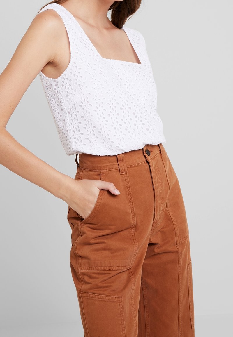 Femme portant un haut blanc sans manches en broderie anglaise et un pantalon taille haute couleur rouille, main dans la poche, sur fond gris.