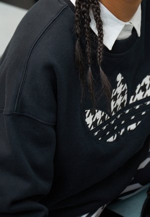 Personne aux cheveux tressés portant un sweat-shirt noir avec un logo à motif pied-de-poule et une chemise blanche à col en dessous.